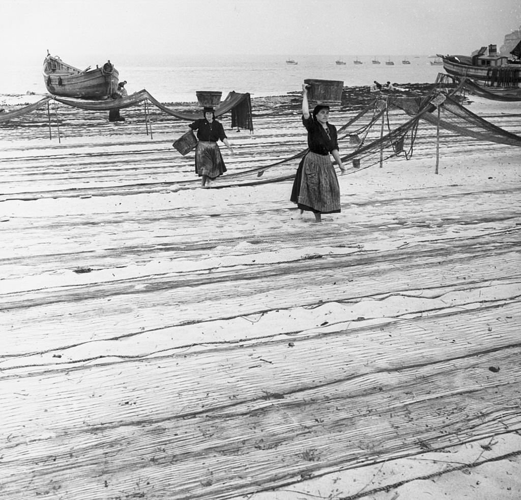 #19 Women carrying fish walk across the drying fishing nets on the beach at Nazare.