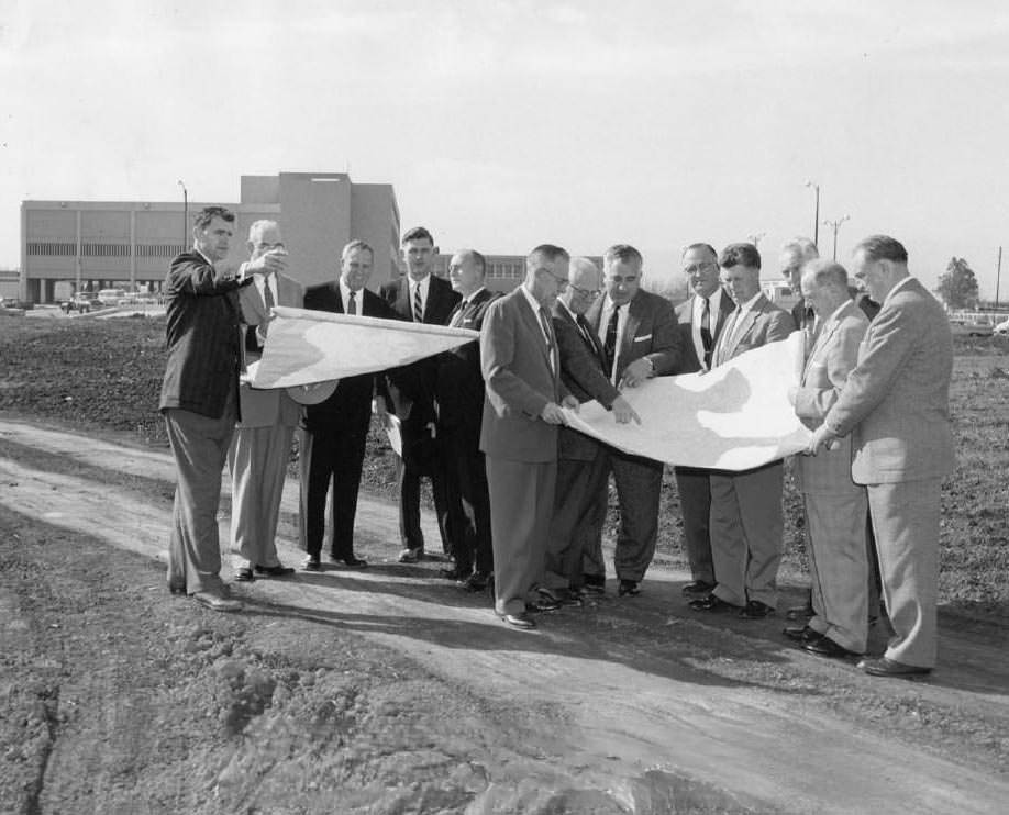 #105 Group of men looking at large plans at construction site, 1959