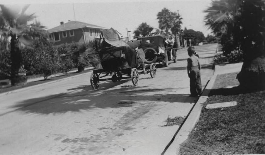 #64 Vehicle towing older car as young boy looks on from the curb, 1932