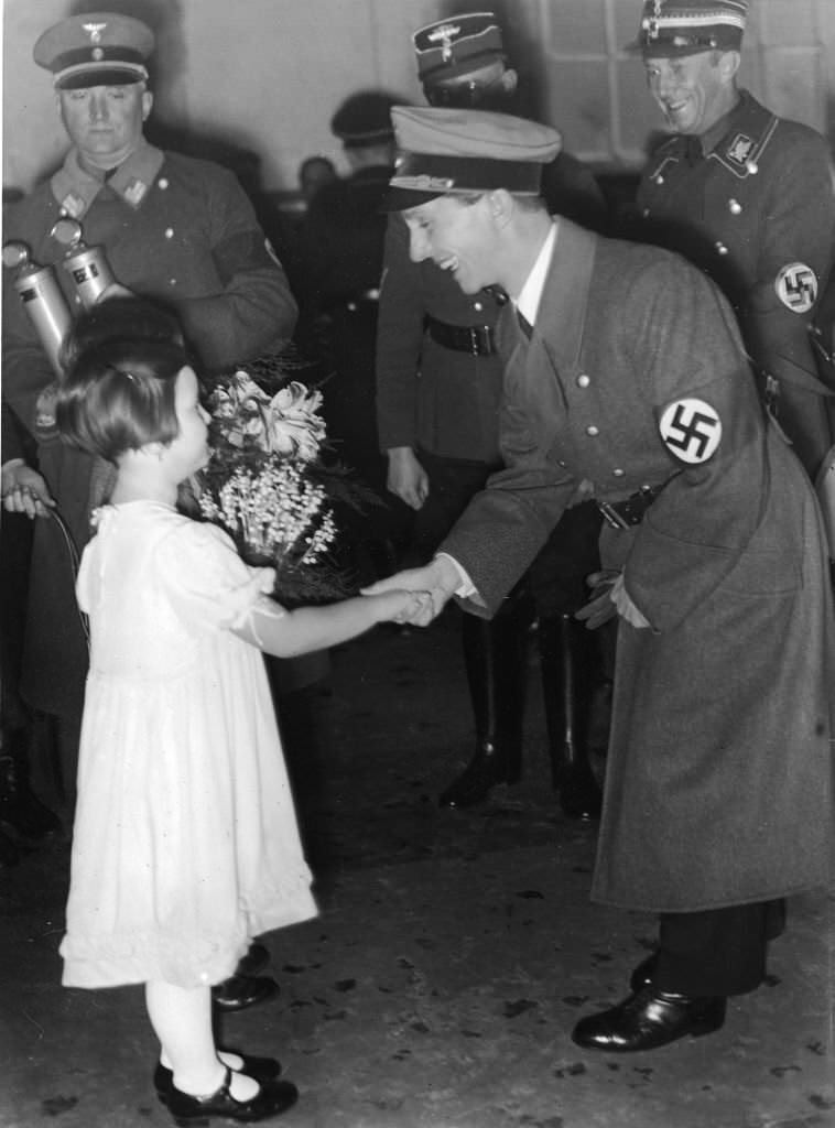 #55 Joseph Goebbels shakes hands with a little girl in a white dress who holds a bouquet of wildflowers, 1930s.