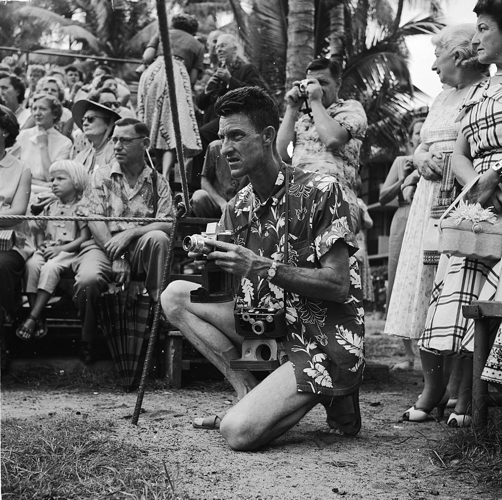 #133 A tourist wearing the traditional Hawaiian shirt kneels with his camera at the ready, awaiting the arrival of the Hula dancers on Honolulu’s Waikiki Beach, in Hawaii, 1953