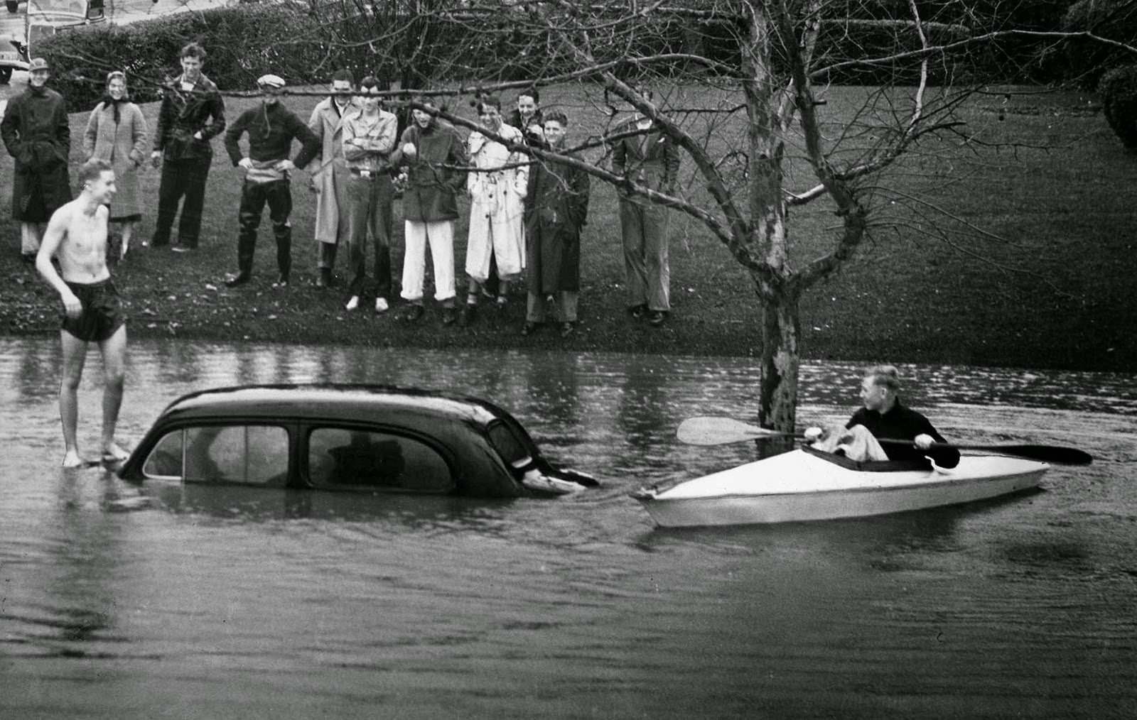 #28 Hugh Beckly stands atop a car almost covered by water at 6th Street near June. Arden Day cruises by in a skiff, 1938