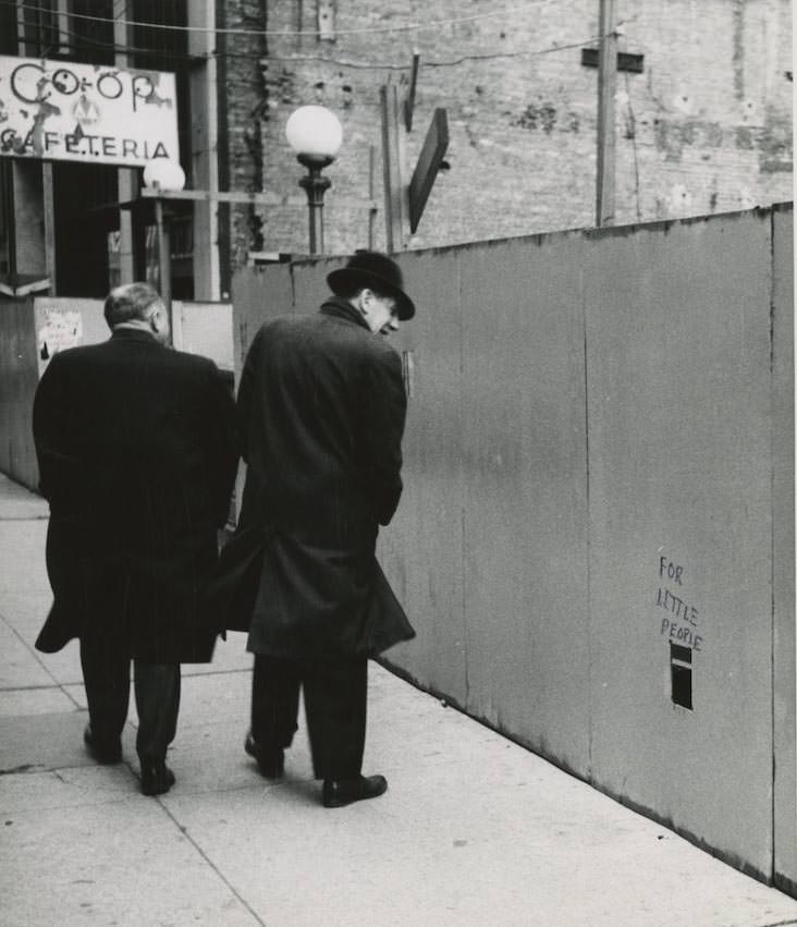#31 Two men walk by a wooden wall with a rectangular cutout that says FOR LITTLE PEOPLE on the street in New York City. Inscription on verso- For Sidewalk Superintendents.