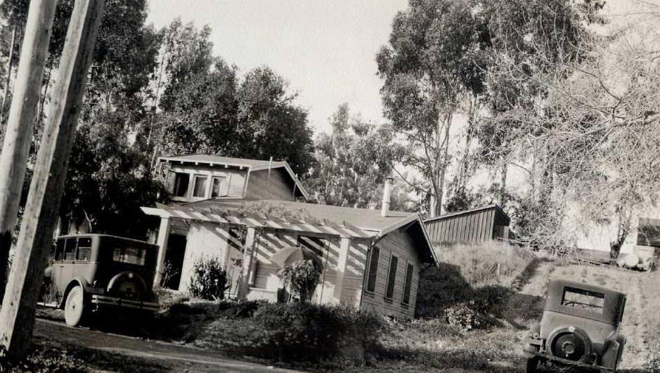 #50 Small house nestled up against wooded hillside. Two cars are parked alongside the dirt roads, 1920s