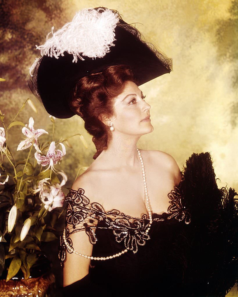 Ava Gardner wearing an off-the-shoulder dress with lace trim, and a broad-brimmed feathered hat, in a promotional portrait for ’55 Days at Peking’, 1963