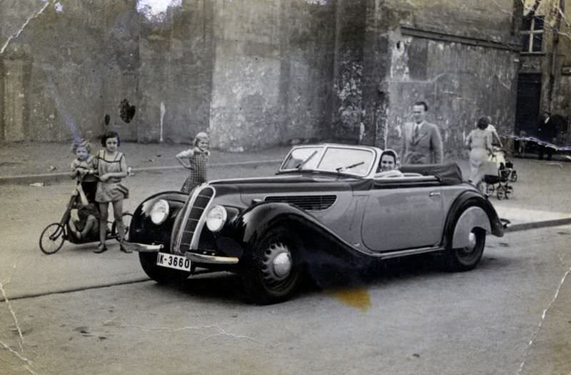 A cheerful lady posing in the driver’s seat of a BMW 327 Cabriolet in a rundown urban backstreet, 1938.