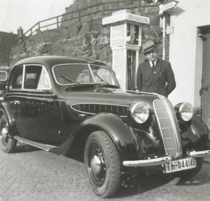 A fellow wearing a double-breasted pinstripe suit and hat posing with a BMW 320 Limousine 2 Türen next to a ‘Bevaulin’ petrol pump, 1939.
