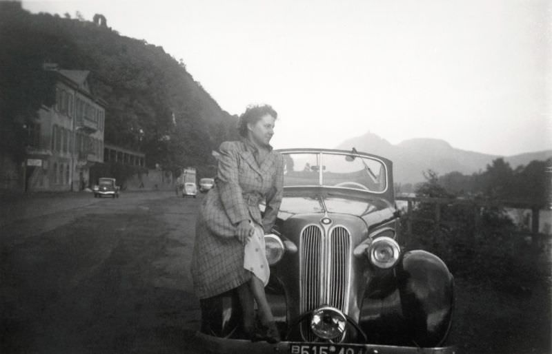 A young lady in a checkered coat posing on the fender a BMW 320 Cabriolet on the side of the road on a bleak summer’s day, 1950.