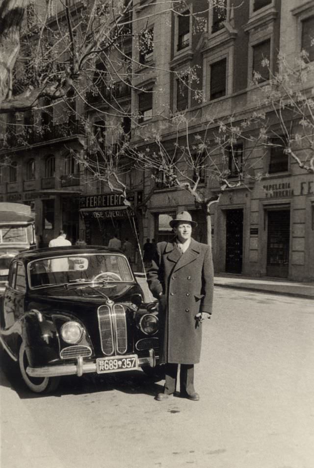 A fellow in a double-breasted coat posing with a BMW 501 on a sunny winter’s day, 1954.