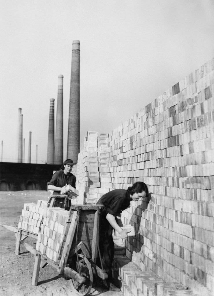 #112 Smoke stack chimneys of the London Brick Company form the backdrop to two women war workers stacking new bricks for the construction industry and the war effort on 1st September 1942 at Stewartby in Bedfordshire, United Kingdom.
