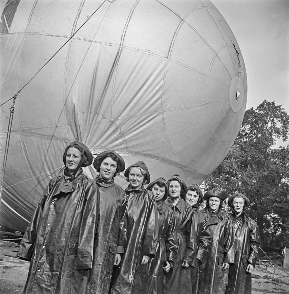 #141 Members of the Women’s Royal Air Force (WRAF) from RAF Balloon Command stand together in their wet weather waterproof oilskins in front of their allocated barrage balloon tethered in Grosvenor Gardens, London. 11th September 1941.