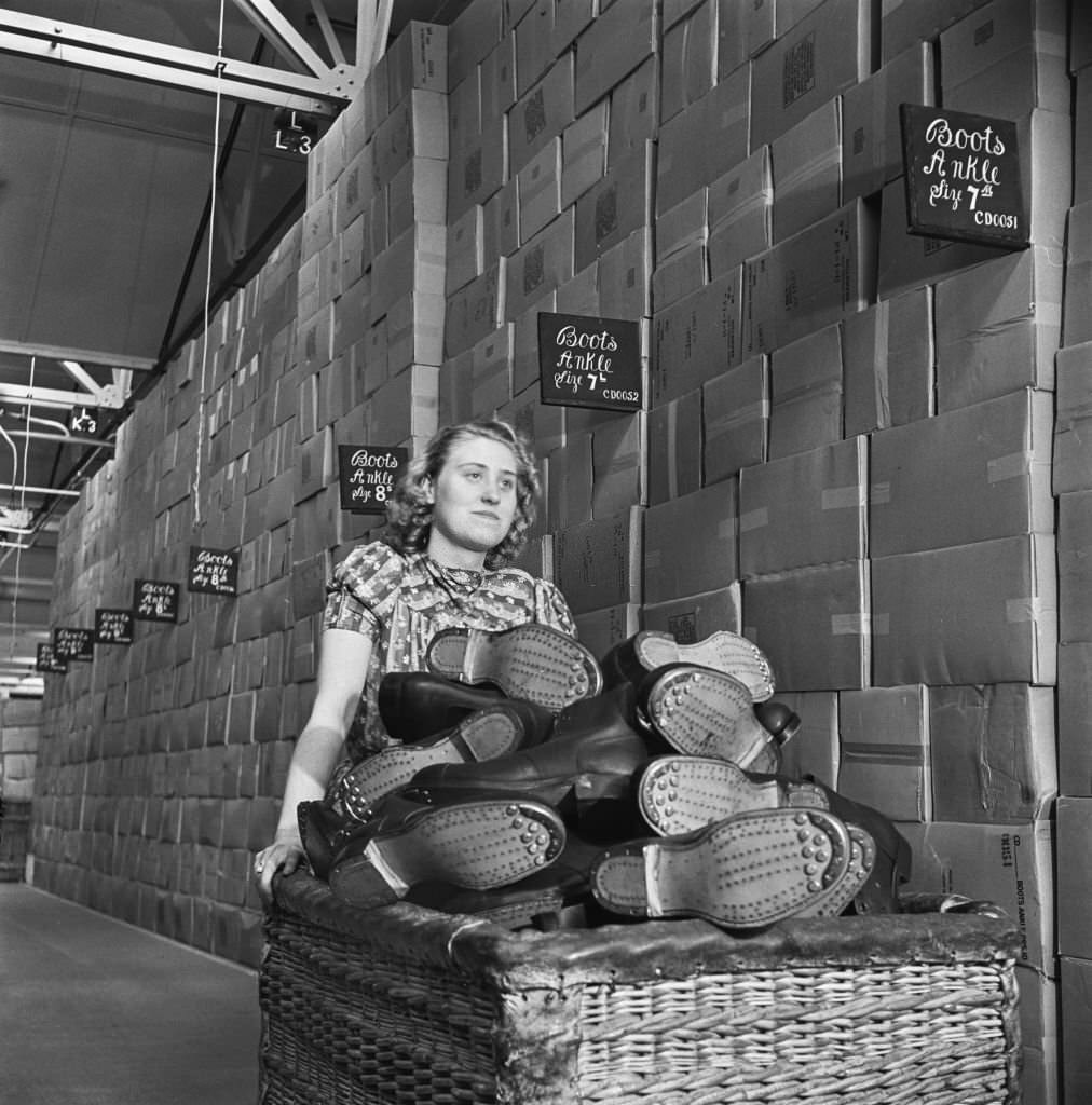 #150 A female warehouse worker pushes a wicker basket full of Britsh Army standard issue Ammunition boots past stacks of hobnail boot boxes at a Central Ordnance Depot for British Army uniform and equipment in England during World War II.