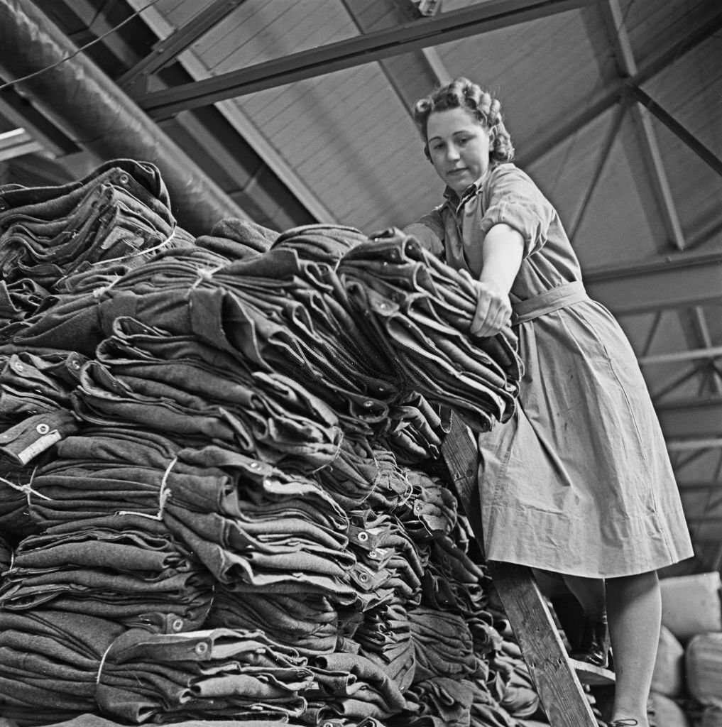 #152 Female warehouse worker Dorothy Durber handles bundles of khaki standard issue military uniform at a Central Ordnance Depot for British Army uniform and equipment in England, 1941.