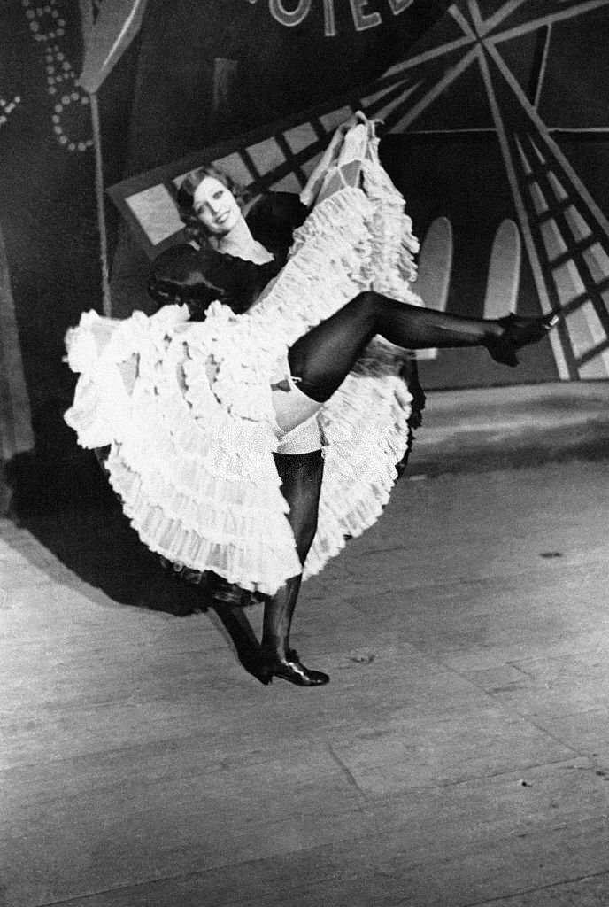 #19 A Cancan dancer takes a break by raising her leg on stage in front of the wings of a windmill, 1900