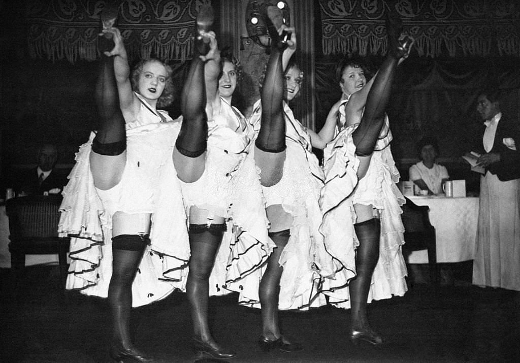 #20 Four cancan dancers raise their legs high in garters and holding each other by the heel under the gaze of spectators and a waiter, 1900