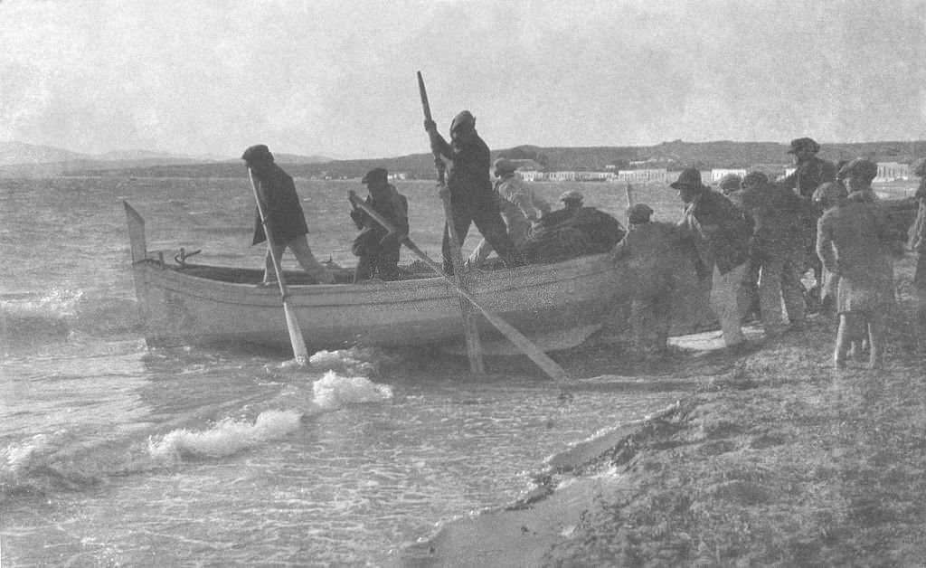 #17 Group of men in a fishing boat holding oars as their boat is pushed into the sea from the shoreline by another group of men as children watch on, Egypt, 1910.