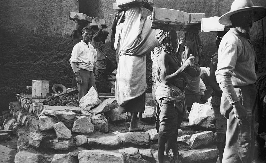 #2 An Archaeologist stands among rocks and stones of an archaeological dig as a group of tribal workers carry boxes on their heads from the site Egypt, 1910.