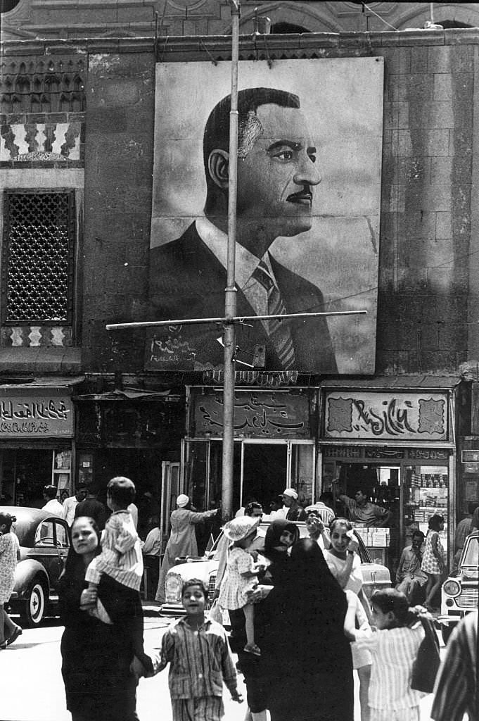 #72 Under a large banner that depicts Egyptian President Gamal Abdel Nasser, pedestrians, some with children, walk on a city street, Cairo, Egypt, 1970.