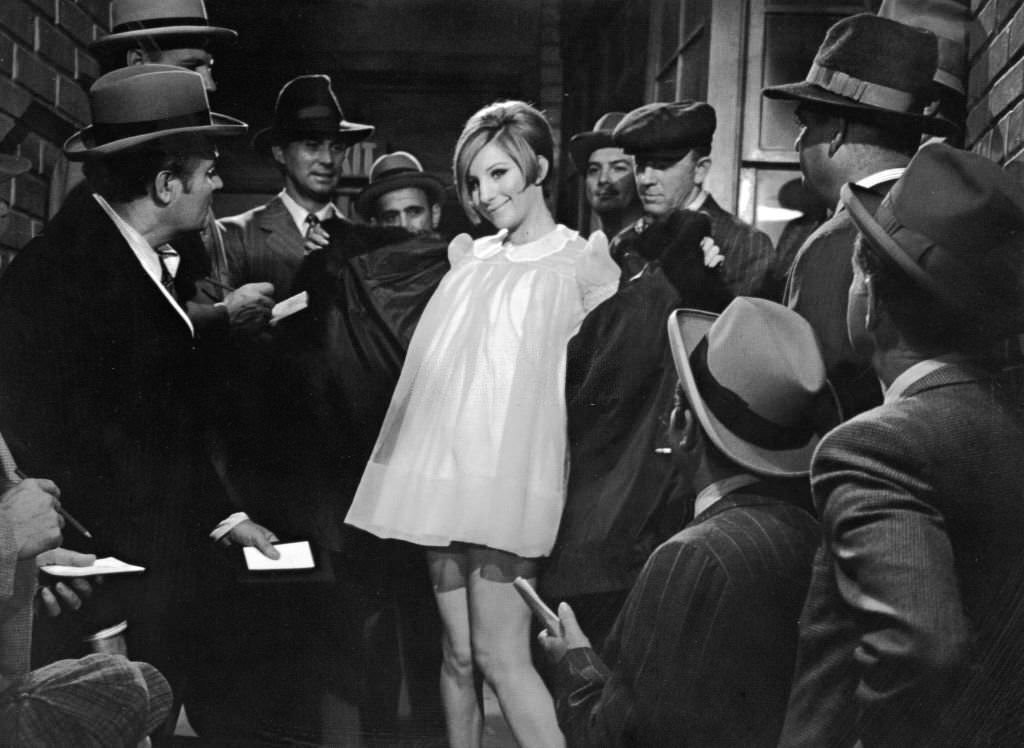 Barbra Streisand opens her coat to reveal a yellow nightgown as reporters gather around her in a still from the film ‘Funny Girl’ 1968