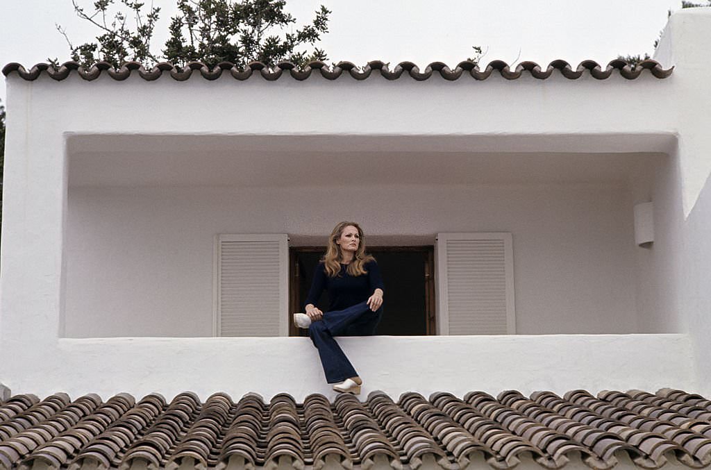 #54 Actress Ursula Andress, in jeans and a dark sweater, sitting on the low wall of her villa’s balcony, Ibiza, 1970s