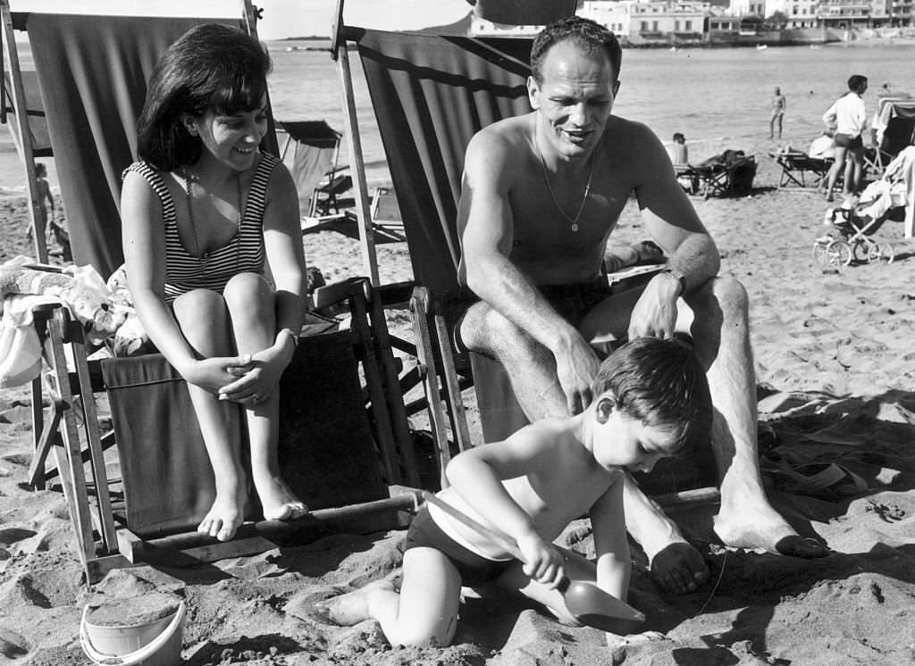 #38 British boxing champion, Henry Cooper sitting on a beach at Las Palmas de Gran Canaria, Gran Canaria with his wife and son, 1965