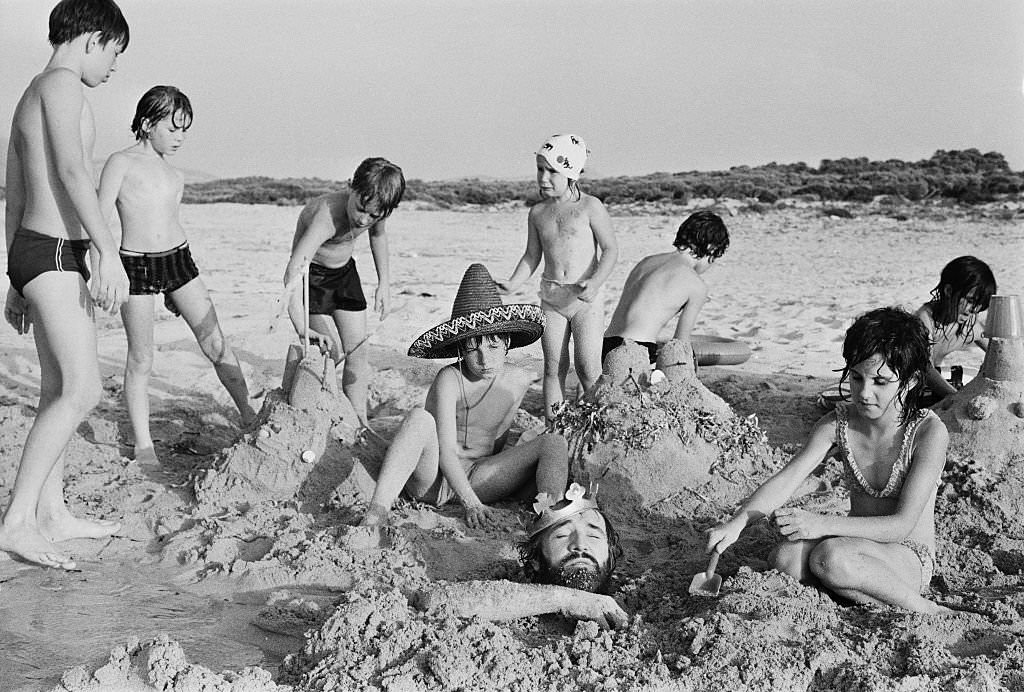 #21 Irish actor Richard Harrisbeing buried in sand by a group of children, whilst on holiday in Majorca, 18th August 1971
