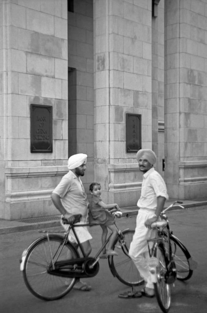#56 Two Asian men resting on their bicycles chatting in the street. Singapore, 1962