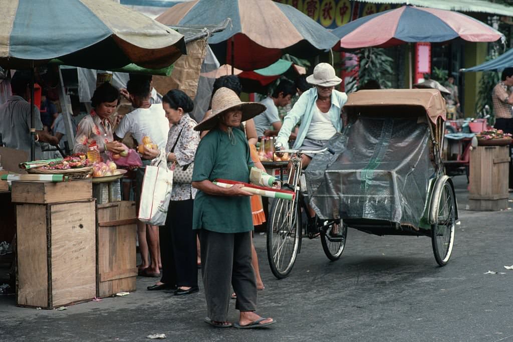 #68 A woman stands next to a man parking a bicycle rickshaw at a market in Singapore.