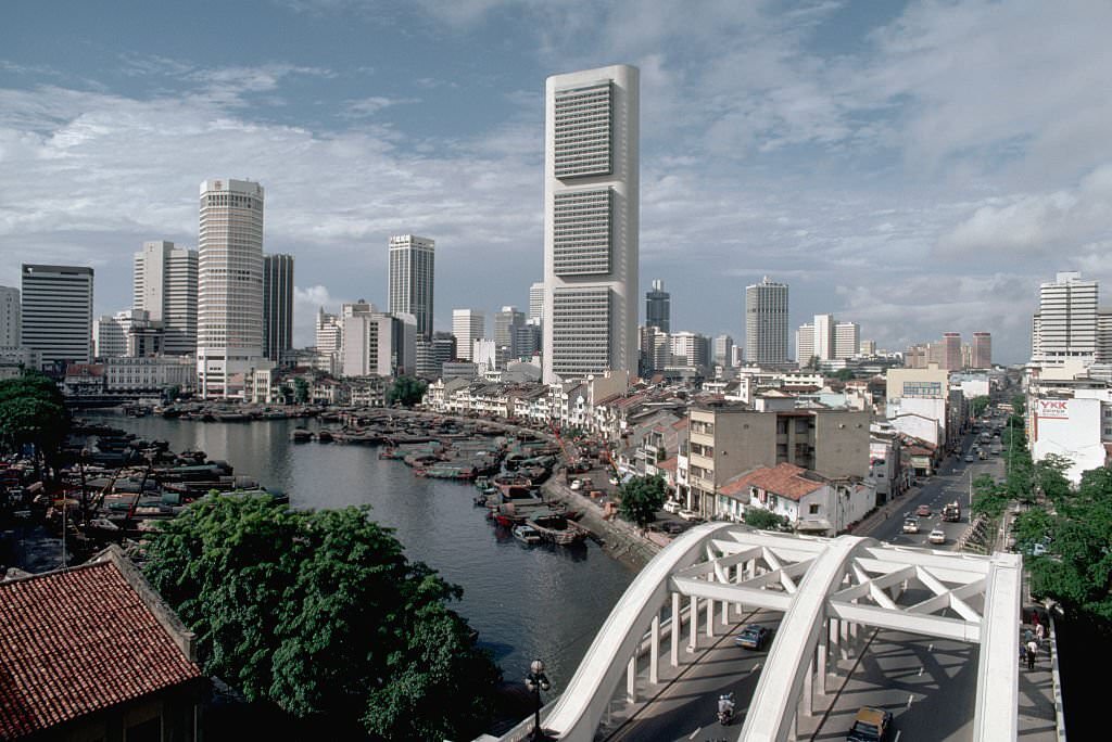 #70 Boats are docked at a quay in the Singapore River near a bridge and a group of skyscrapers, 1980s