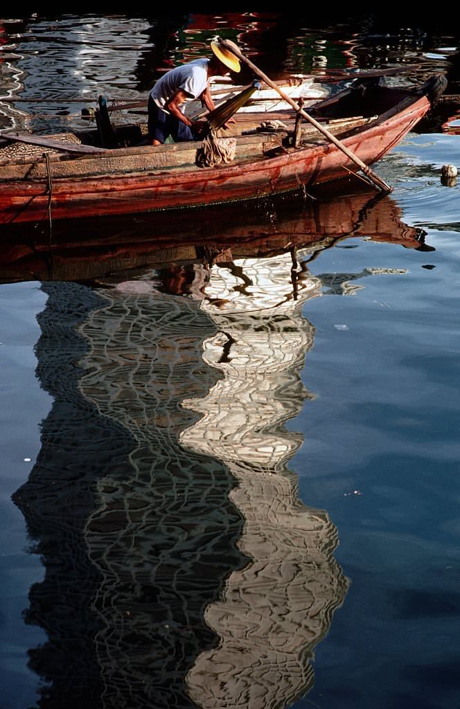 #75 A fisherman prepares his boat by a reflection of a skyscraper in Singapore, 1980s