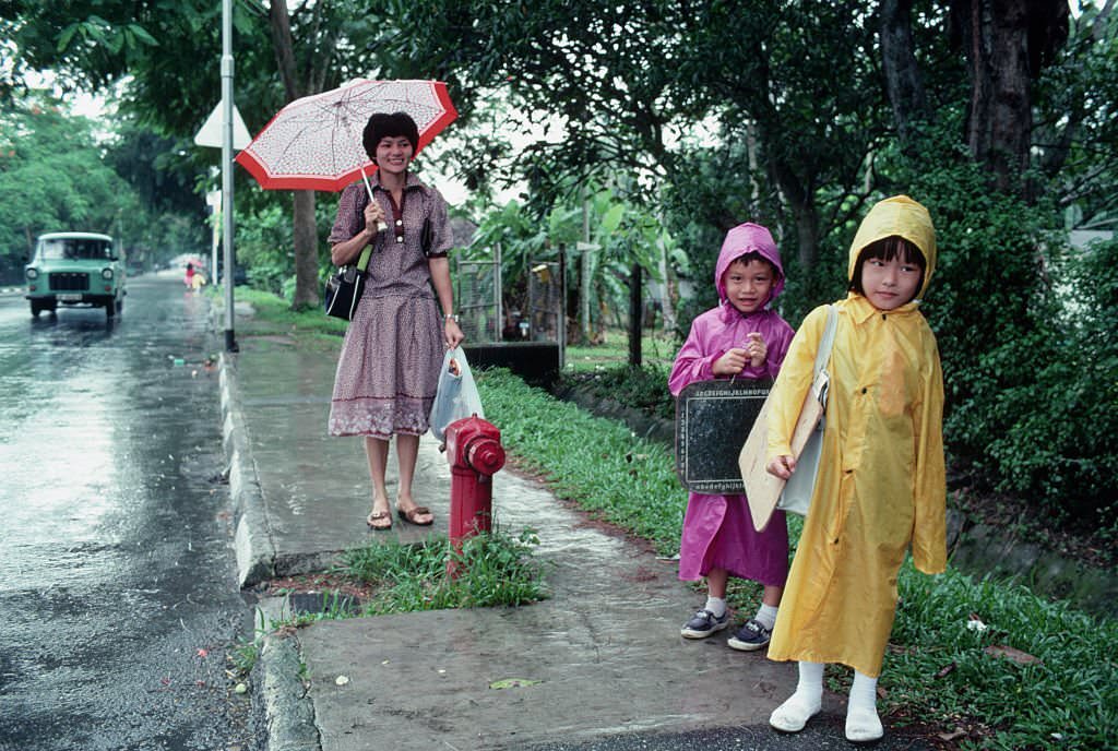 #77 Two school children wait at a bus stop in Singapore, accompanied by a woman who is possibly their mother, 1980s