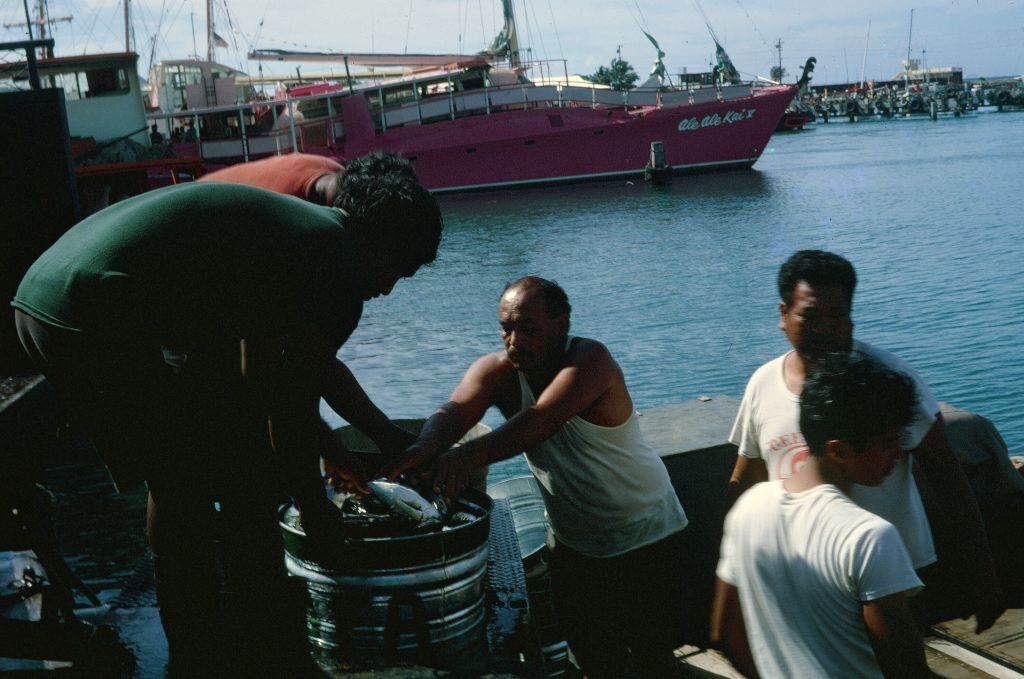 #12 Native fishermen load their catch into a barrel, with the Ale Ale Kai IV catamaran visible in the background, Honolulu, Hawaii, 1970.