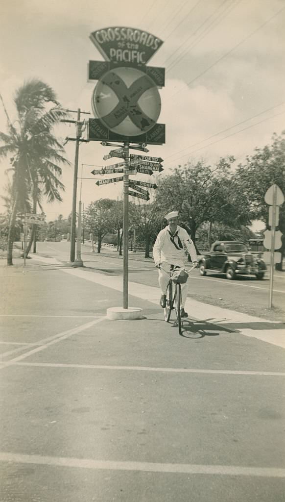 #85 Sailor rides bicycle by Crossroads of the Pacific sign, 1940s