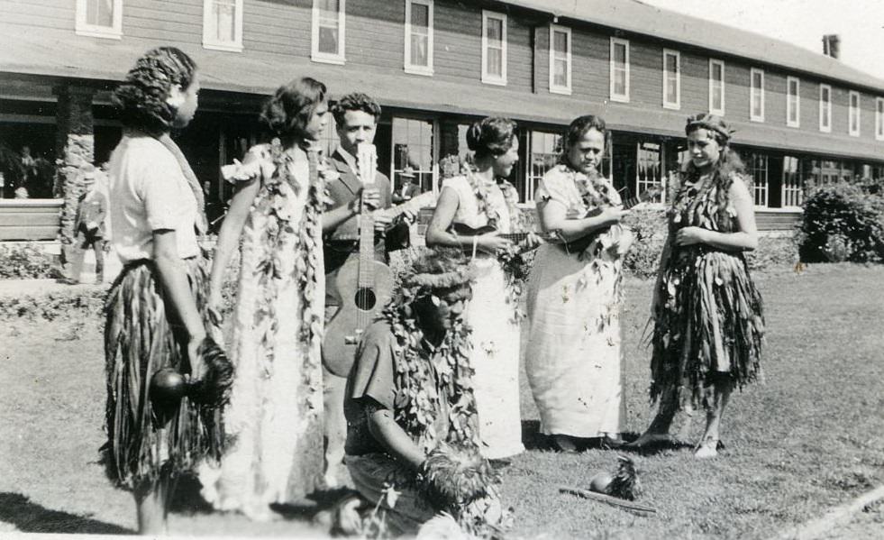 #104 Women wearing leis and hula skirts standing on grass in front of building holding musical instruments, 1940s