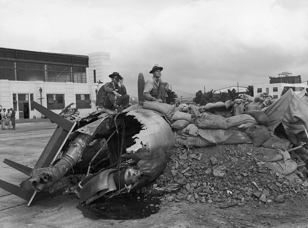 #137 Two servicemen sit on the wreckage of a bomber, surrounded by dirt and sandbags, on Hickam Field after the Japanese attack on Pearl Harbor, Honolulu, Hawaii, 1941