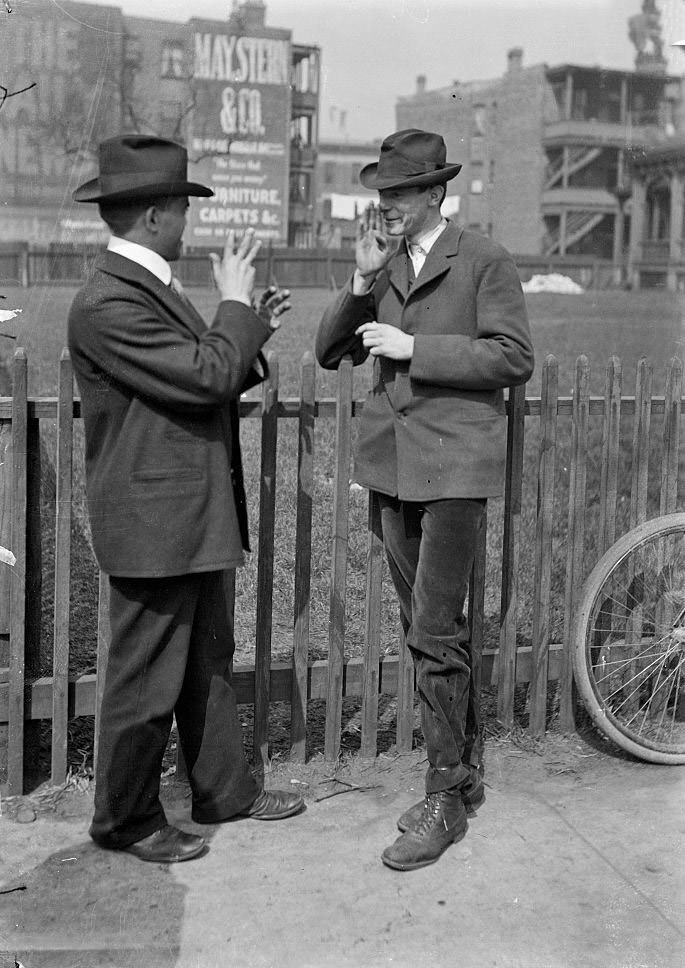 #139 Two deaf men standing in front of a fenced yard or field signing to each other during the Automatic Electric Telephone Company strike, Chicago, Illinois, April 23, 1903.