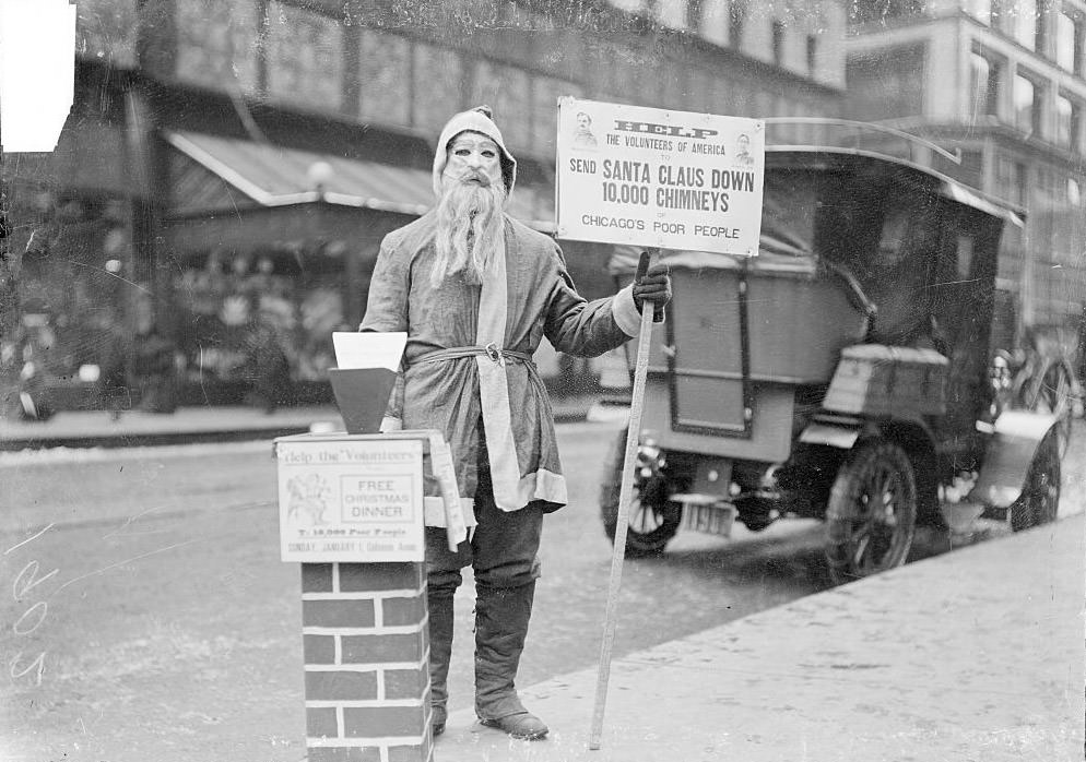 #167 A man dressed up as Santa Claus holding a sign that reads, Help the Volunteers of America to send Santa down 10,000 chimneys of Chicago’s poor people, on the sidewalk of a commercial street, Chicago, 1902