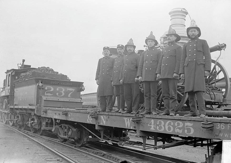 #216 Six firemen standing in front of a fire engine on a train flatcar, 1900s.