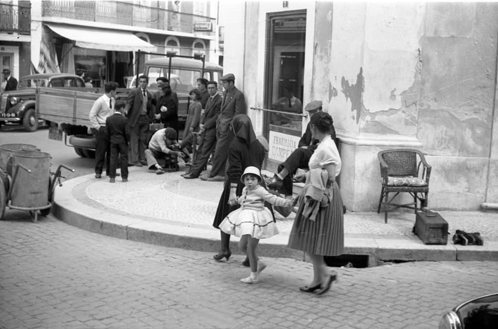 #84 Street scene People in front of a shop entrance. A little girl, wears a skirt and a hat, is hold by two women