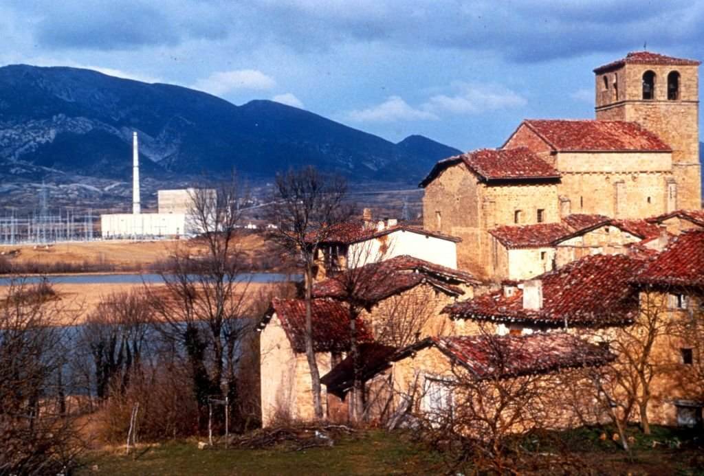 #40 Mid shot of medieval stone buildings and leafless trees, with the Ebro river, Santa Maria de Garona Nuclear Power Plant, and mountains in the background, Burgos, Spain, 1970.
