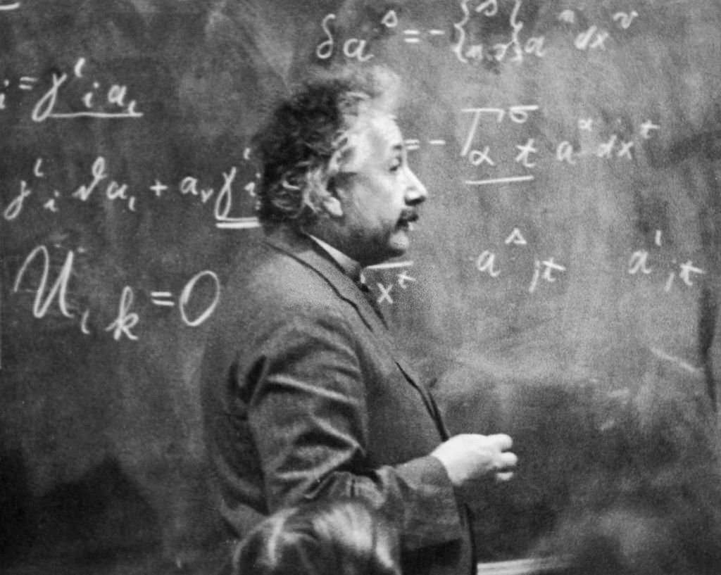 #46 Albert Einstein standing beside a blackboard with chalk-marked mathematical calculations written across it, 1931