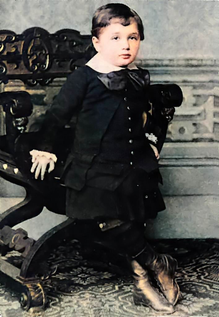 #17 Three-year-old Albert Einstein, at approximately three years old, wearing a black suit and large neck bow, leaning against an X-chair, and facing the camera with a calm expression, 1882.