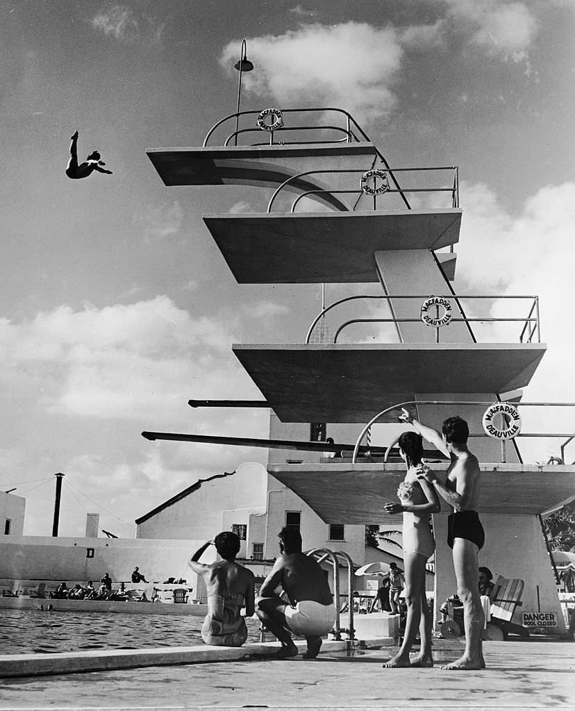 #119 People sit poolside and watch as a swimmer dives off the high platform of the Olympic-size swimming pool at the MacFadden – Deauville Hotel, 1950s