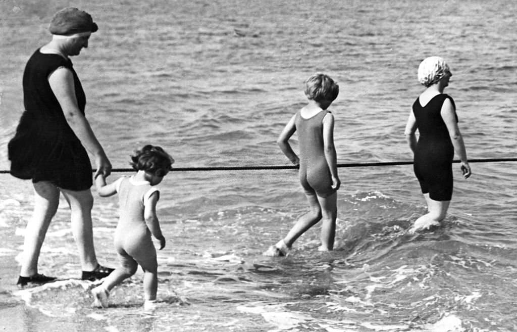 #137 A woman and her children wearing one-piece swimming-suit secure themselves holding a rope as they enter in the water, 1937