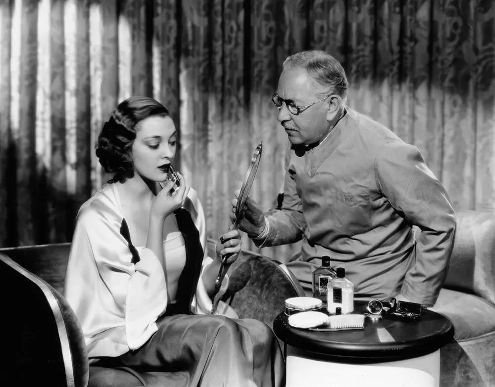 #10 Actress Kathleen Burke applies lipstick with the help of Factor. 1930.