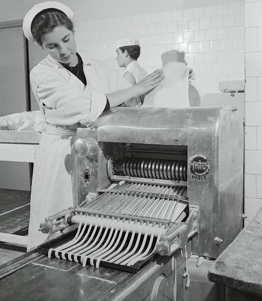 #33 Woman Feeding Dough into a Machine. Passing through mechanical channels, the future bread sticks resemble thick spaghetti as they begin to take on an elongated appearance.