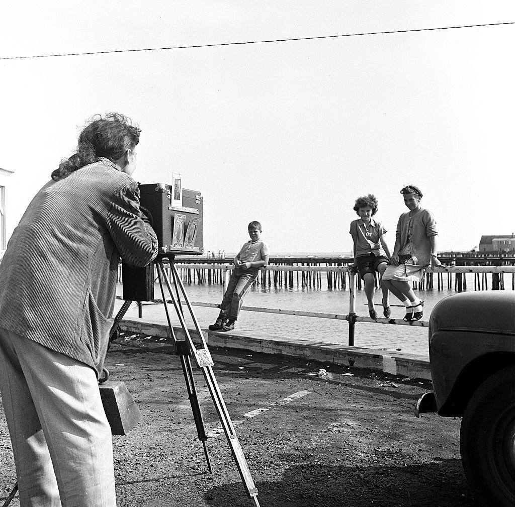 #45 hree visitors sit or lean on the pier’s railings as a photographer uses a camera on a tripod to take portraits, Provincetown, Massachusetts, 1948.