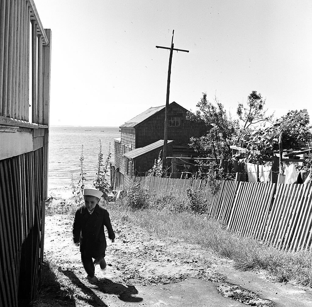 #75 A young boy wearing a sailor’s hat walks past the deck of a seaside house on Cape Cod, Provincetown, Massachussetts, 1947.