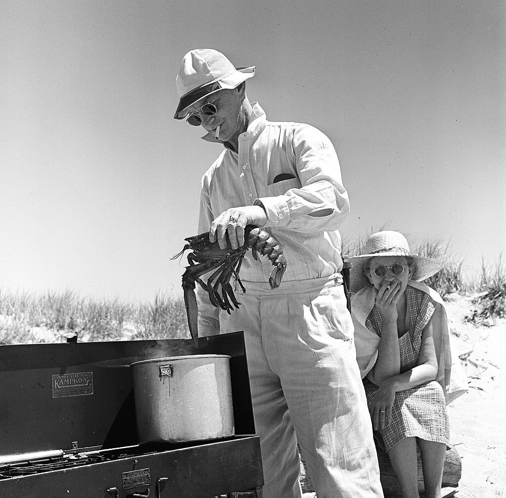 #77 A man prepares to cook a lobster in a pot set up on an outdoor grill, with a woman seated behind him on Cape Cod, Provincetown, Massachussetts, 1947.