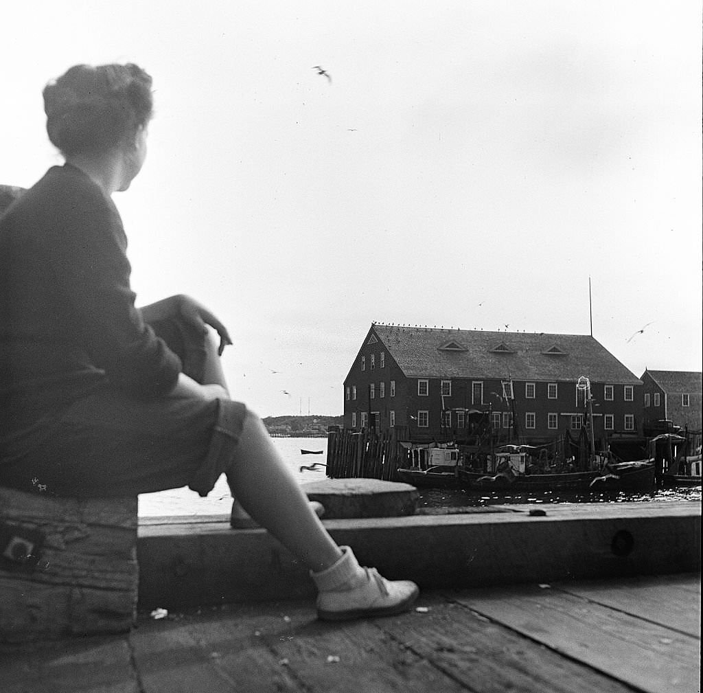 #80 A young woman sitting on the pier looks out onto some dockside buildings on Cape Cod, Provincetown, Massachussetts, 1947.